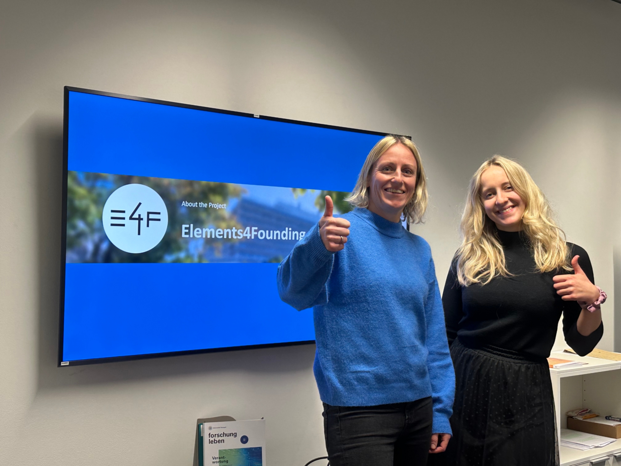 Foto der wissenschaftlichen Mitarbeiterinnen Kathrin Lichius (links) und Mia Zsohár (rechts) vor einem Bildschirm, der eine Präsentationsfolie in den Farben der Universität mit dem Logo des Projektes Elements4Founding zeigt.