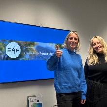 Foto der wissenschaftlichen Mitarbeiterinnen Kathrin Lichius (links) und Mia Zsohár (rechts) vor einem Bildschirm, der eine Präsentationsfolie in den Farben der Universität mit dem Logo des Projektes Elements4Founding zeigt.